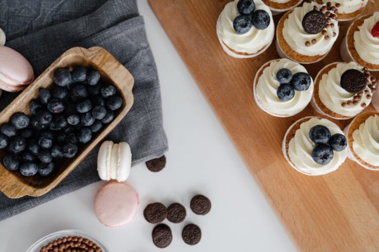 Top view of homemade cupcakes served on wooden tray near chocolate sweets and macaroons with blueberries in wooden container